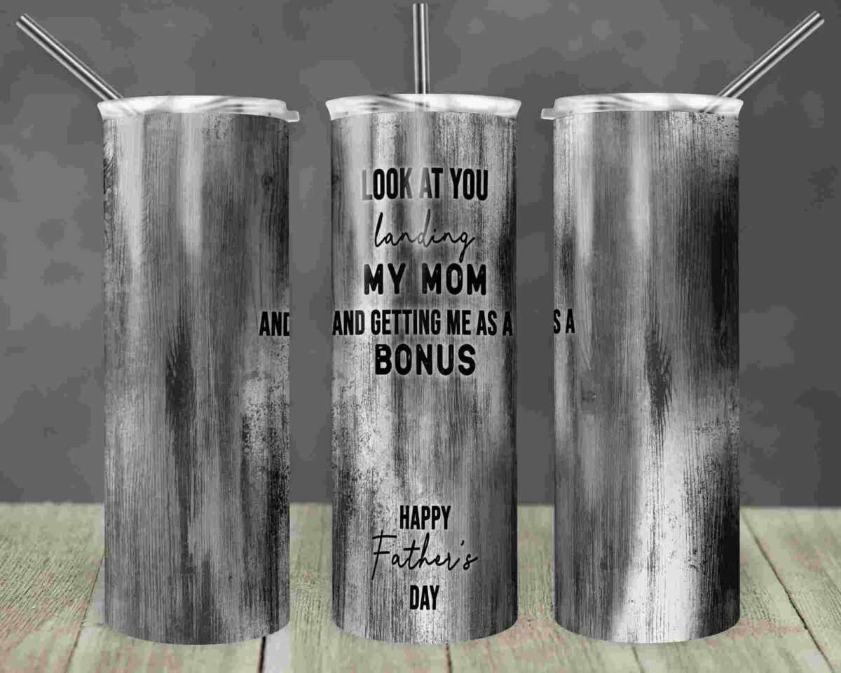 A grayscale tumbler with the text: "Look at you landing my mom and getting me as a bonus. Happy Father's Day." The tumbler has three views displayed on a wooden surface.