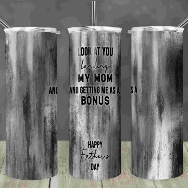 A grayscale tumbler with the text: "Look at you landing my mom and getting me as a bonus. Happy Father's Day." The tumbler has three views displayed on a wooden surface.