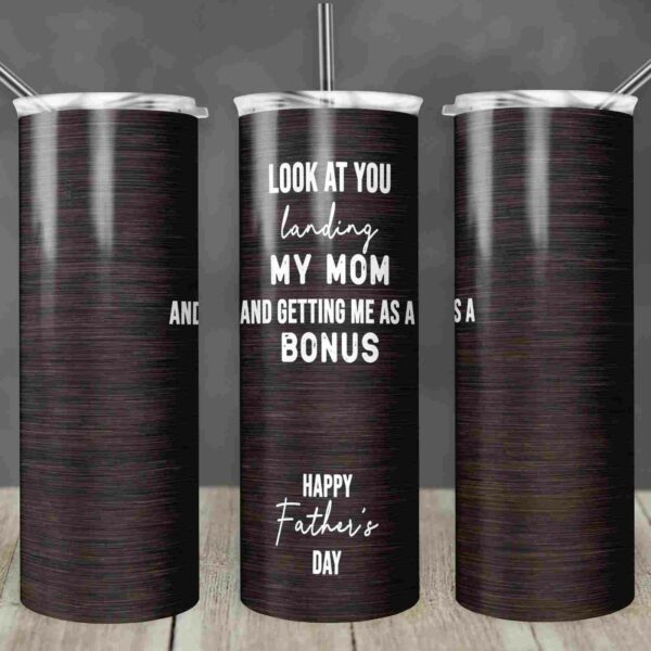 A dark wood-textured tumbler with a silver lid and straw is shown from three angles. The text on the tumbler reads, "Look at you landing my mom and getting me as a bonus. Happy Father's Day." The background is blurred and rustic.