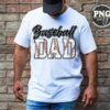 A bearded man wearing a white T-shirt that reads "Baseball Dad" stands in front of a brick wall. The word "Baseball" is styled in a cursive font, while "Dad" is in a bold, block font with red stitching resembling a baseball. The man has a dark cap and blue jeans.