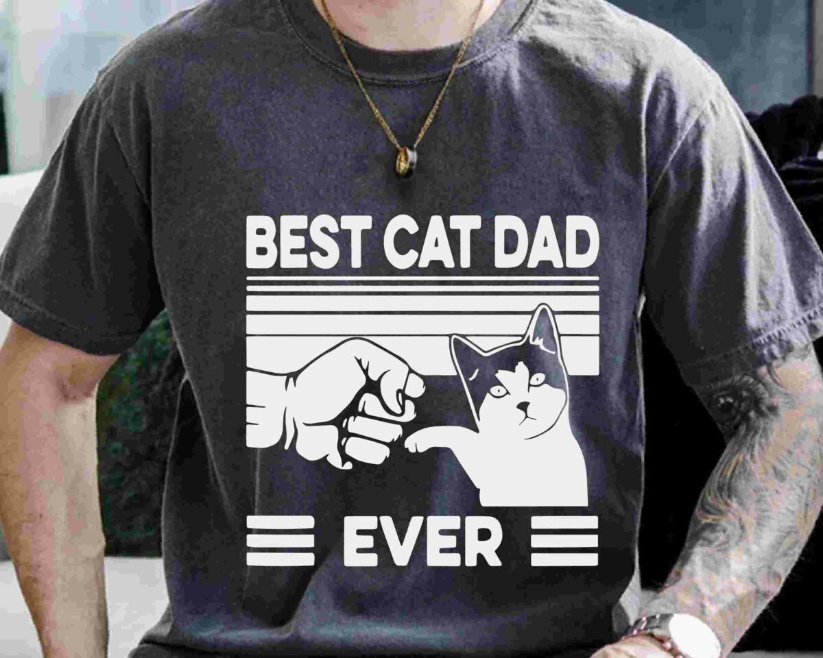 A person is wearing a dark gray t-shirt with the text "BEST CAT DAD EVER" and an illustration of a fist bumping a cat's paw.