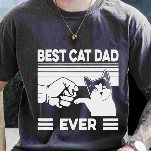 A person is wearing a dark gray t-shirt with the text "BEST CAT DAD EVER" and an illustration of a fist bumping a cat's paw.