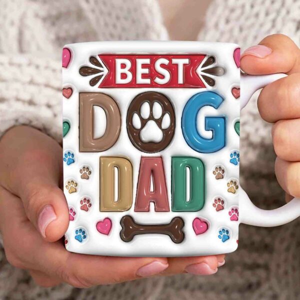 A person holds a white mug with colorful text that reads "Best Dog Dad," featuring paw prints, hearts, and a bone design.