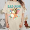 Alt Text: A person wearing a beige t-shirt featuring a cartoon dog and the text "RAD MOM." The person is standing in a room with patterned decor and a clay vase.