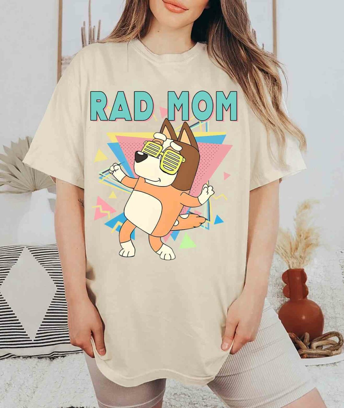 Alt Text: A person wearing a beige t-shirt featuring a cartoon dog and the text "RAD MOM." The person is standing in a room with patterned decor and a clay vase.