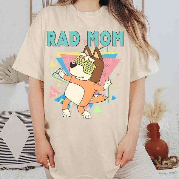 Alt Text: A person wearing a beige t-shirt featuring a cartoon dog and the text "RAD MOM." The person is standing in a room with patterned decor and a clay vase.