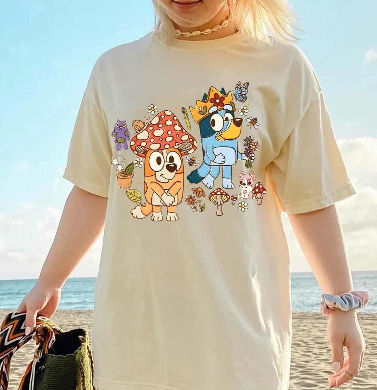 Person wearing a beige t-shirt with cartoon dog characters and mushrooms, standing near the beach holding a woven bag.