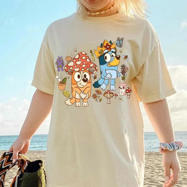 Person wearing a beige t-shirt with cartoon dog characters and mushrooms, standing near the beach holding a woven bag.