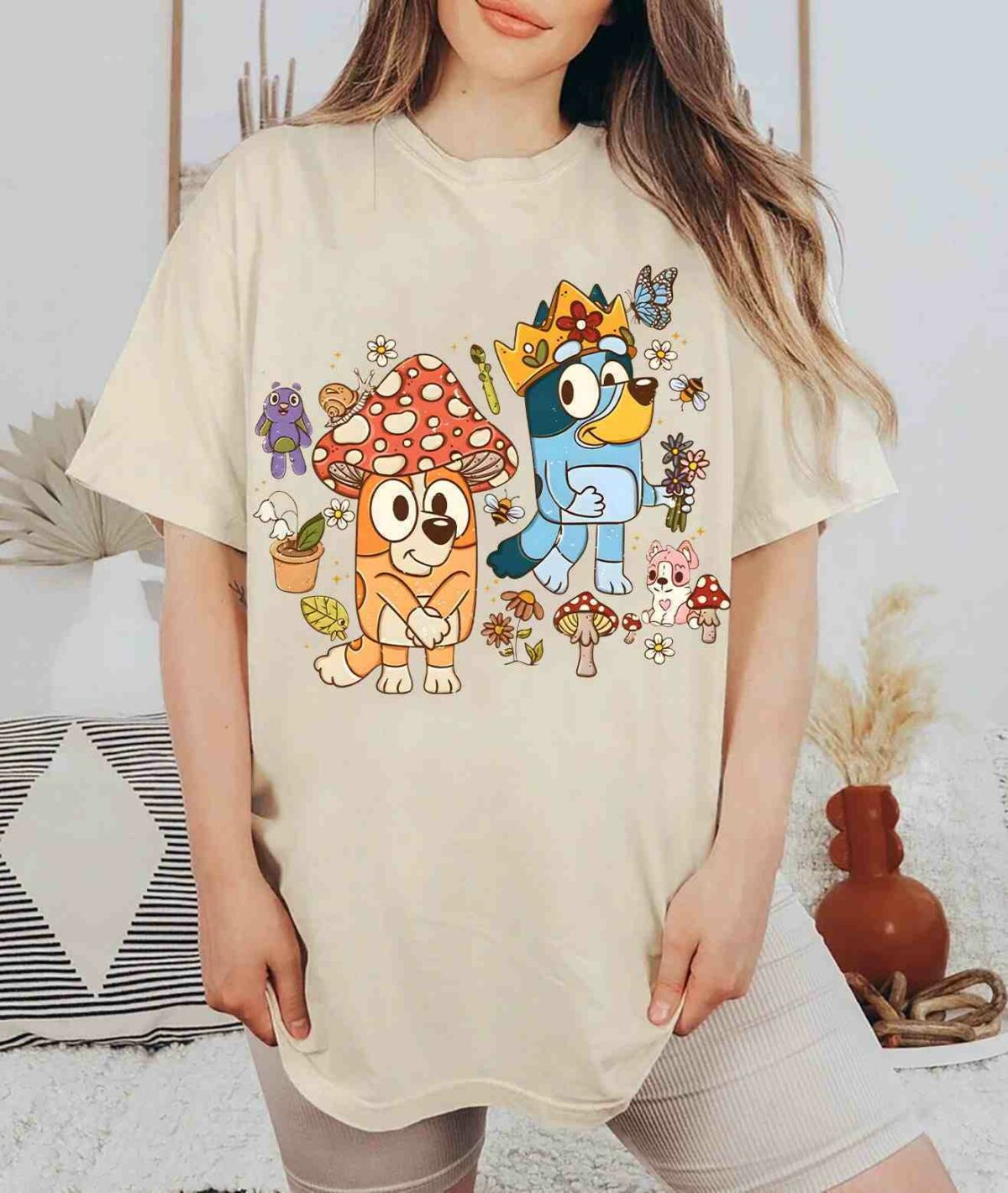 Person wearing a beige t-shirt featuring colorful cartoon characters, including a blue dog with a crown and an orange dog with a mushroom hat, standing amid various flora and fauna.