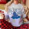 A person wearing a light gray sweatshirt with a cute cartoon narwhal saying, "Bye, Buddy! Hope you find your dad!" They are also holding a white and red mug with a Santa face, sitting cross-legged in red and black plaid pants. Holiday decor is visible in the background.