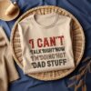 A beige T-shirt with the text "I CAN'T TALK RIGHT NOW I'M DOING HOT DAD STUFF" lies neatly folded in a wicker basket on a dark blue fabric background, with a spool of thread and beige cloth nearby.