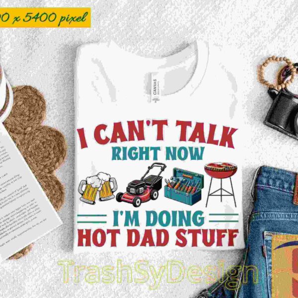 Alt Text: White T-shirt with text: "I CAN'T TALK RIGHT NOW I'M DOING HOT DAD STUFF" alongside images of a beer, grill, and lawnmower. Laid out with jeans, a camera, a book, and sunglasses.