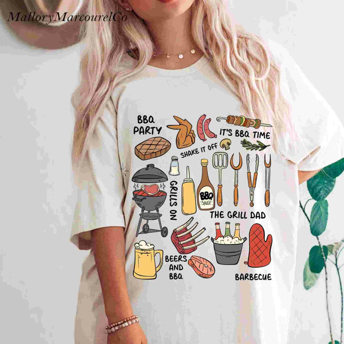 A person wearing a white t-shirt with colorful illustrations related to barbecue. The drawings include a grill, various barbecue foods, condiments, and utensils. Text phrases such as "BBQ PARTY," "THE GRILL DAD," "BEERS AND BBQ," and "BARBECUE" are also displayed.