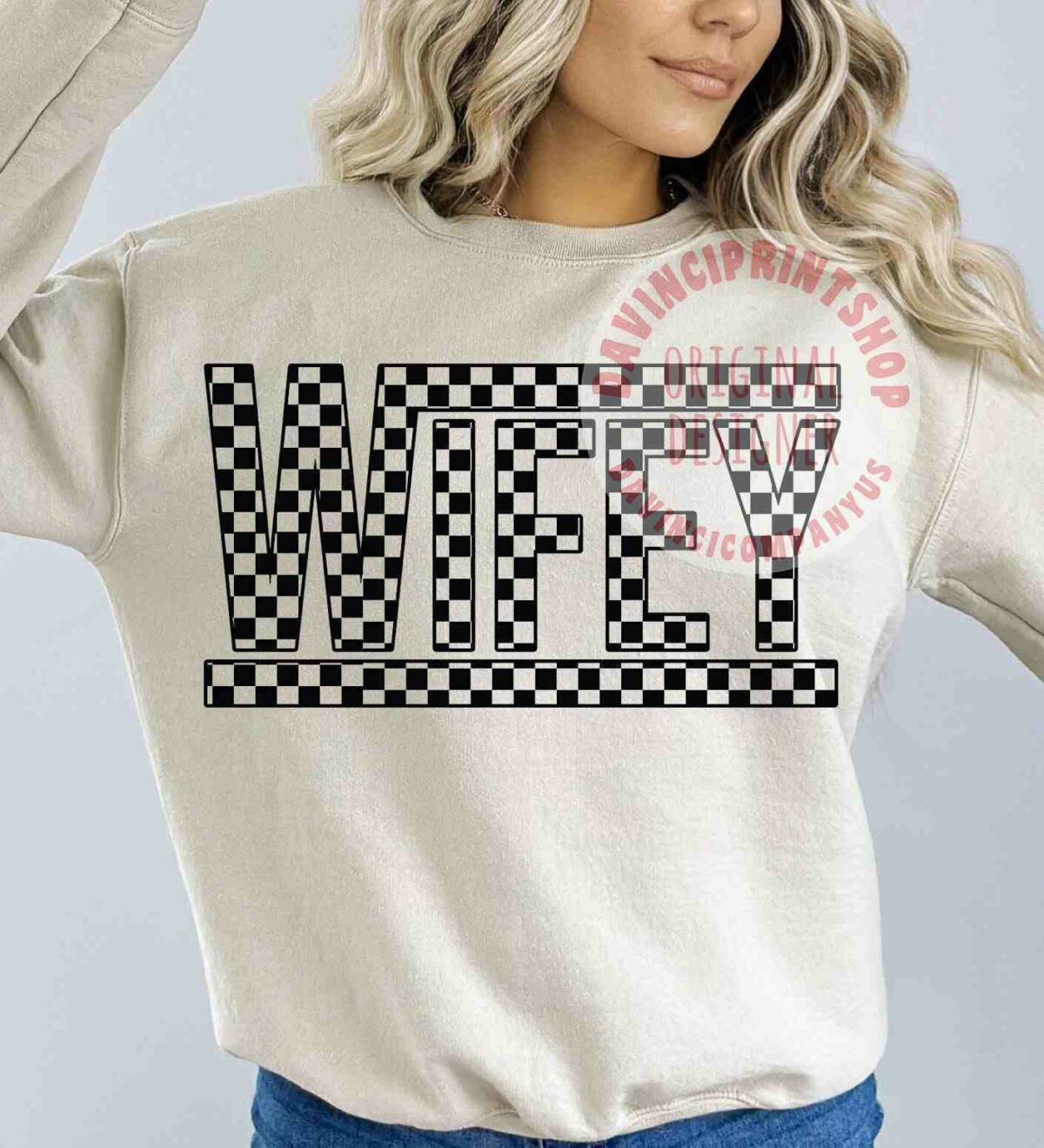 Image of a woman wearing a beige sweatshirt with the word "WIFEY" in bold, checkered black and white letters across the chest. She has long, wavy blonde hair and is posing with one hand raised to her head. The watermark says "VINCI PRINT SHOP ORIGINAL DESIGN" in red text.