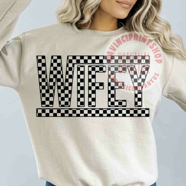 Image of a woman wearing a beige sweatshirt with the word "WIFEY" in bold, checkered black and white letters across the chest. She has long, wavy blonde hair and is posing with one hand raised to her head. The watermark says "VINCI PRINT SHOP ORIGINAL DESIGN" in red text.