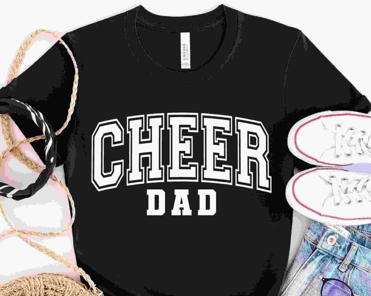 A black T-shirt with the text "CHEER DAD" in bold white letters is laid out flat. Surrounding the shirt are a straw hat, a black and white striped ribbon, a pair of white sneakers with red trim, and a pair of light-wash denim jeans.