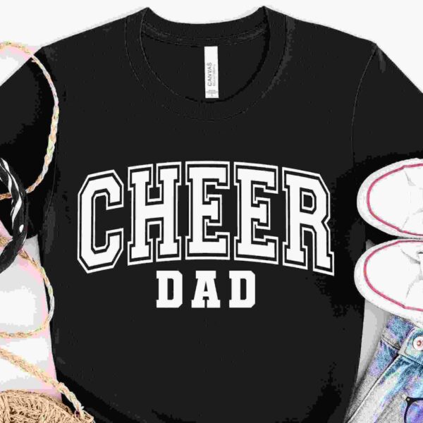 A black T-shirt with the text "CHEER DAD" in bold white letters is laid out flat. Surrounding the shirt are a straw hat, a black and white striped ribbon, a pair of white sneakers with red trim, and a pair of light-wash denim jeans.