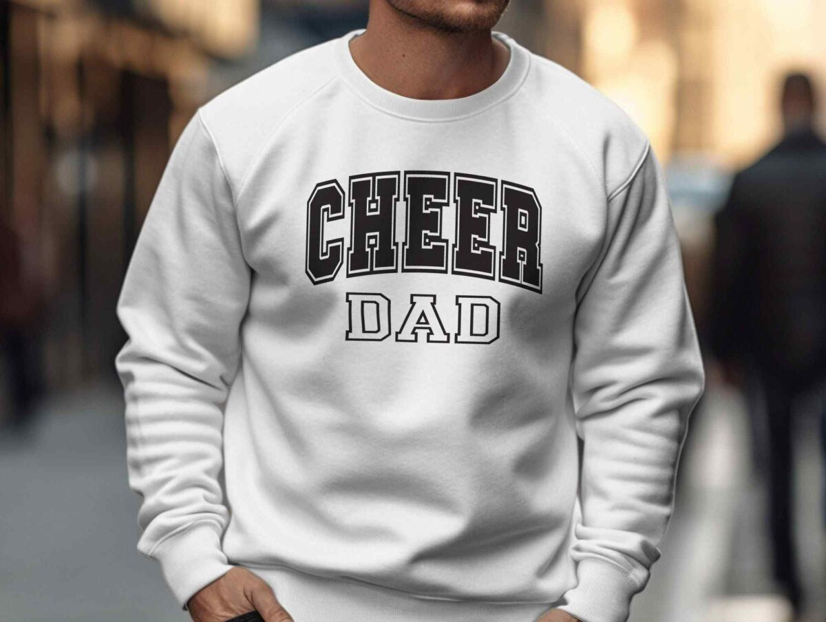 A man wearing a white sweatshirt with the text "CHEER DAD" in bold black letters stands on a city sidewalk. The background shows a blurred cityscape with a person walking away in the distance.