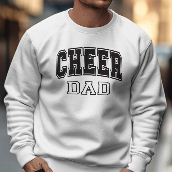 A man wearing a white sweatshirt with the text "CHEER DAD" in bold black letters stands on a city sidewalk. The background shows a blurred cityscape with a person walking away in the distance.