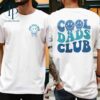 Two people wearing white T-shirts with "Cool Dads Club" printed in blue and green letters on the back and a smaller, similar design on the front with a lightning bolt logo.