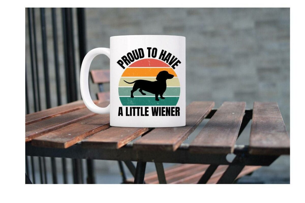 A white mug on a wooden table features a silhouette of a dachshund and the text "Proud to Have a Little Wiener" against a striped background.