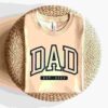 A folded beige T-shirt with the word "DAD" in bold black lettering and "EST. 2023" inscribed below is displayed on a round, woven wicker surface against a white background.