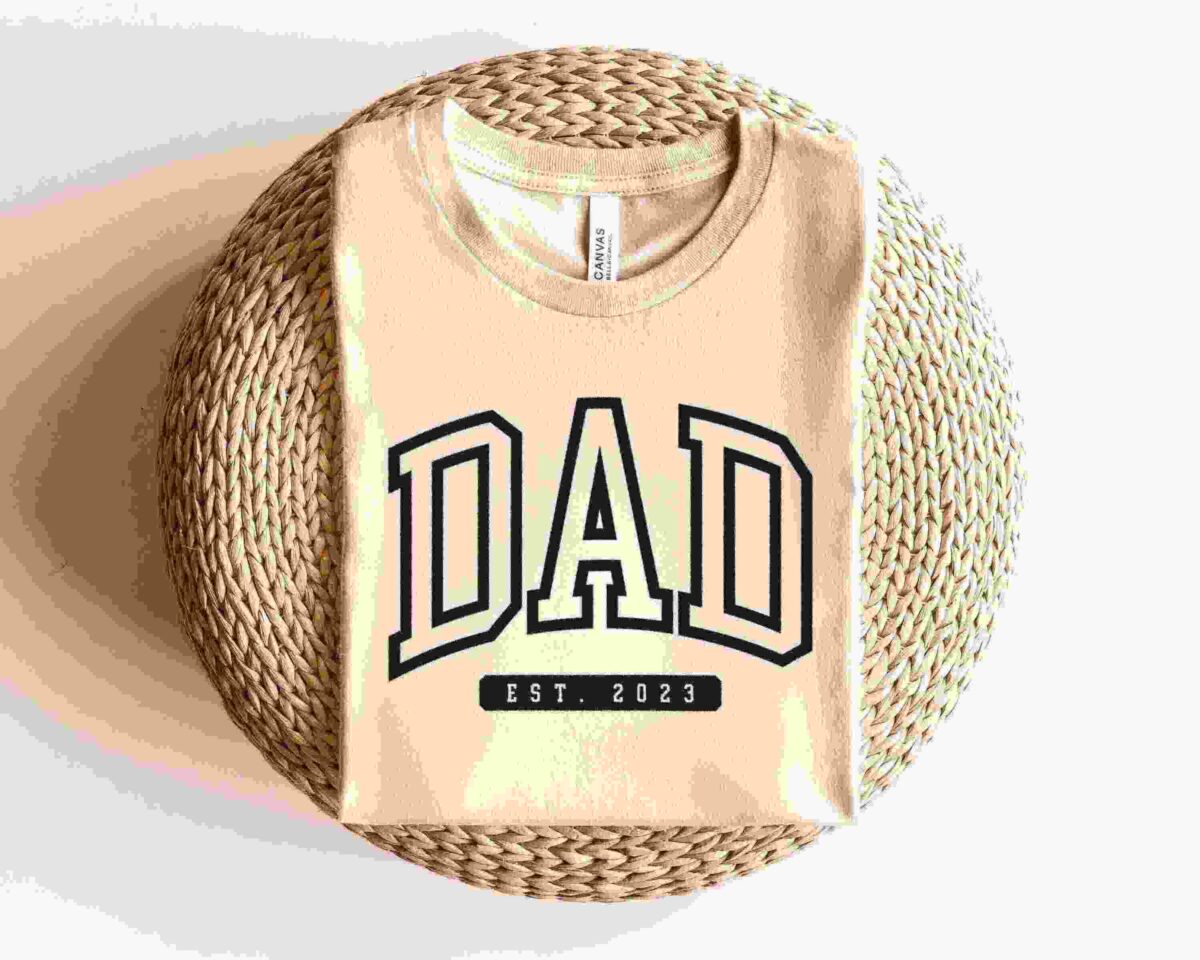 A folded beige T-shirt with the word "DAD" in bold black lettering and "EST. 2023" inscribed below is displayed on a round, woven wicker surface against a white background.