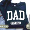 A navy blue sweatshirt featuring the word "DAD" in large white letters, with "EST. 2023" beneath it. The sweatshirt is laid out on a light surface alongside a pair of light blue jeans, a cup of coffee, and a pair of light brown sneakers. The text "SVG & PNG" and "MP" logos are also present.