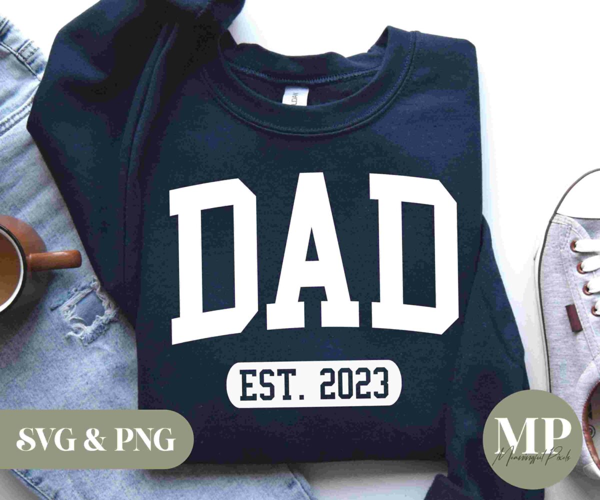 A navy blue sweatshirt featuring the word "DAD" in large white letters, with "EST. 2023" beneath it. The sweatshirt is laid out on a light surface alongside a pair of light blue jeans, a cup of coffee, and a pair of light brown sneakers. The text "SVG & PNG" and "MP" logos are also present.