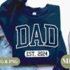 Navy blue sweatshirt with the word "DAD" and "EST. 2024" printed in white on the front, laid on light blue jeans next to a brown mug and sneakers. Text overlay reads "SVG & PNG" and "MP Moving Press".