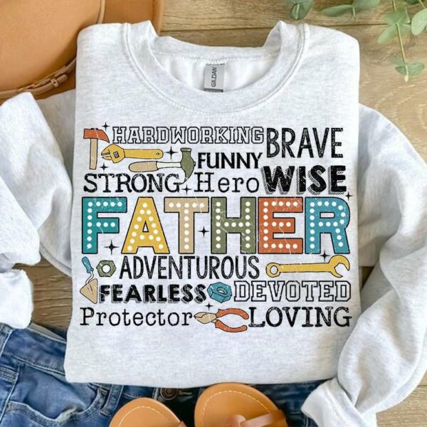 A white sweatshirt displaying colorful words describing a father, including "hardworking," "brave," "funny," "strong," "hero," "wise," "adventurous," "fearless," "devoted," "protector," and "loving." The shirt is laid on a wooden surface next to denim jeans and a leather bag.