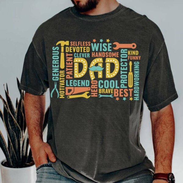 A man is wearing a dark shirt with colorful words and illustrations surrounding the word "Dad" in the center. Words such as "Wise," "Cool," "Patient," "Protector," and "Hero" are interspersed with tool icons, highlighting positive attributes commonly associated with fathers.