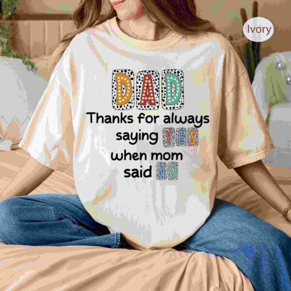 A person is sitting on a bed wearing a white T-shirt with the text, "Dad, Thanks for always saying yes when mom said no.