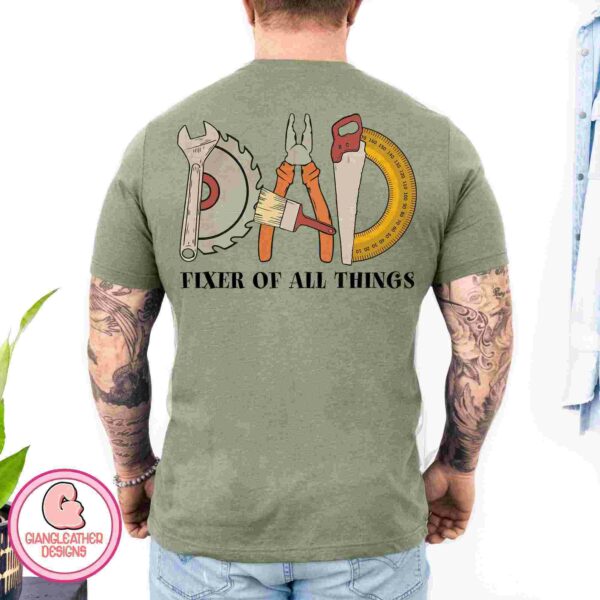 A man with tattoos on his arms wearing a green T-shirt that says "DAD FIXER OF ALL THINGS" with illustrations of tools forming the word "DAD".