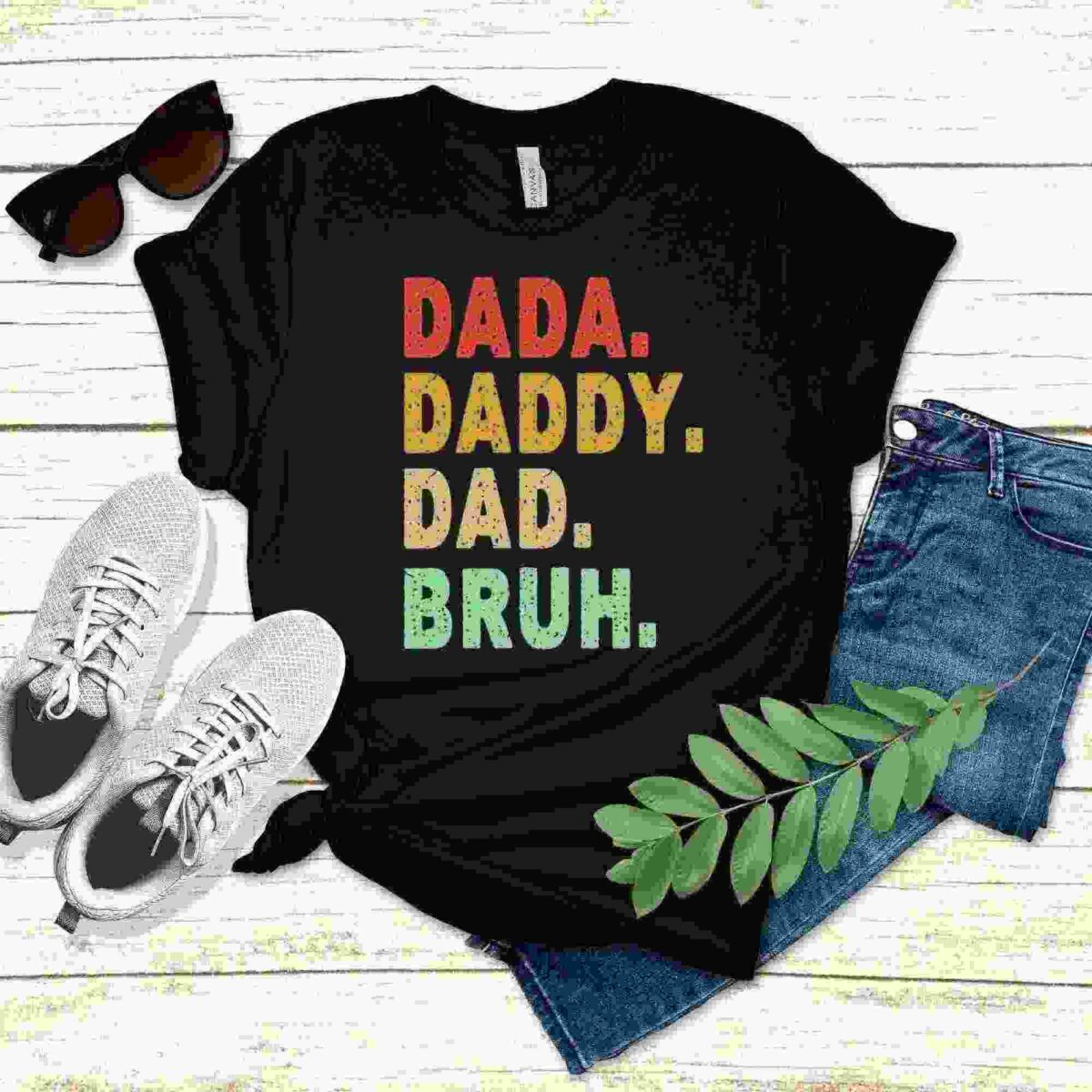 Alt Text: Black T-shirt with the text "DADA. DADDY. DAD. BRUH." in colorful fonts, placed alongside white sneakers, jeans, sunglasses, and a green leaf on a light wooden surface.