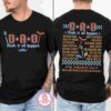 Front and back view of a black T-shirt. The front reads "DAD, Mate it all happen" with various dad-related graphics. The back lists numerous dad activities and is titled "The Fatherhood Tour.