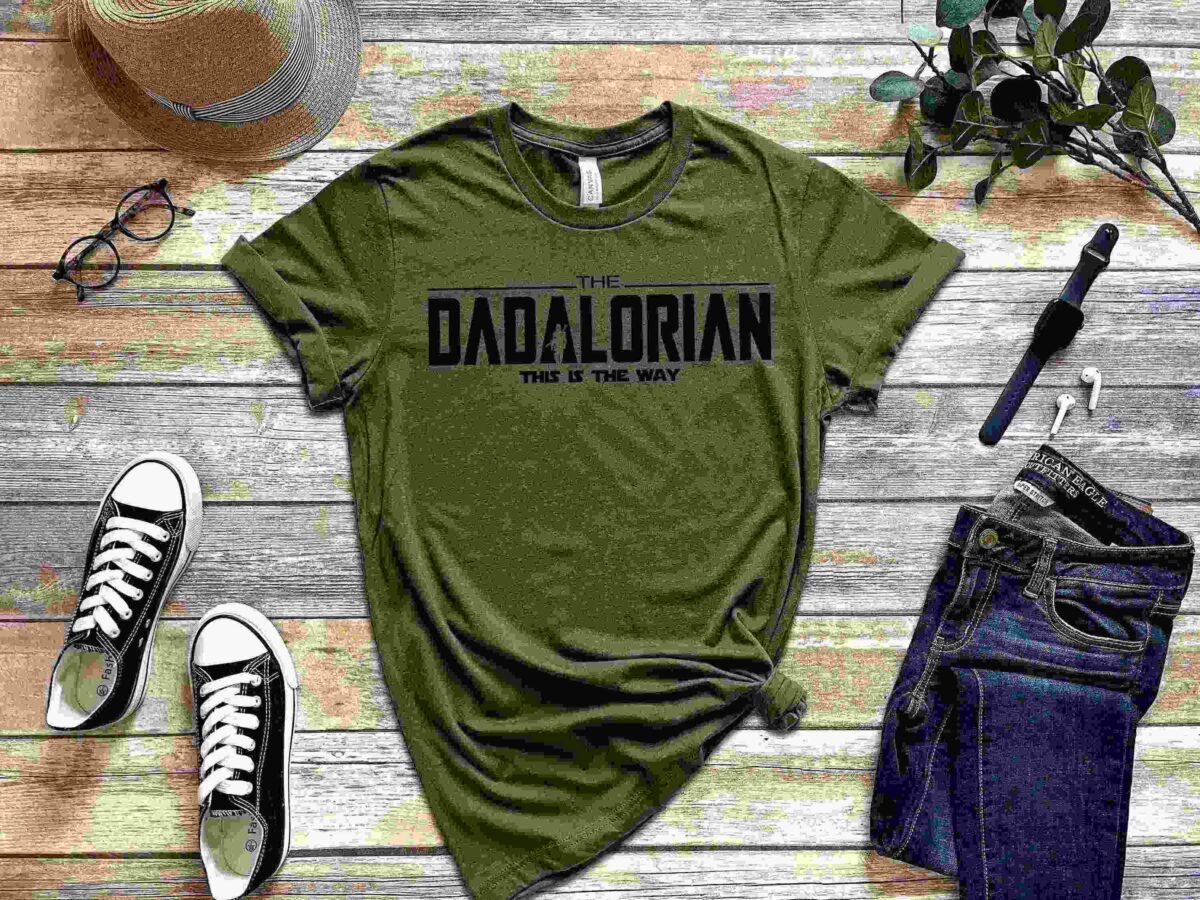 A green t-shirt with "The Dadalorian: This is the Way" printed on it, displayed with a pair of black sneakers, blue jeans, a watch, glasses, and a hat on a wooden surface with a plant.