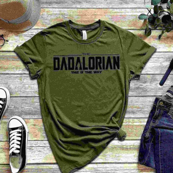 A green t-shirt with "The Dadalorian: This is the Way" printed on it, displayed with a pair of black sneakers, blue jeans, a watch, glasses, and a hat on a wooden surface with a plant.