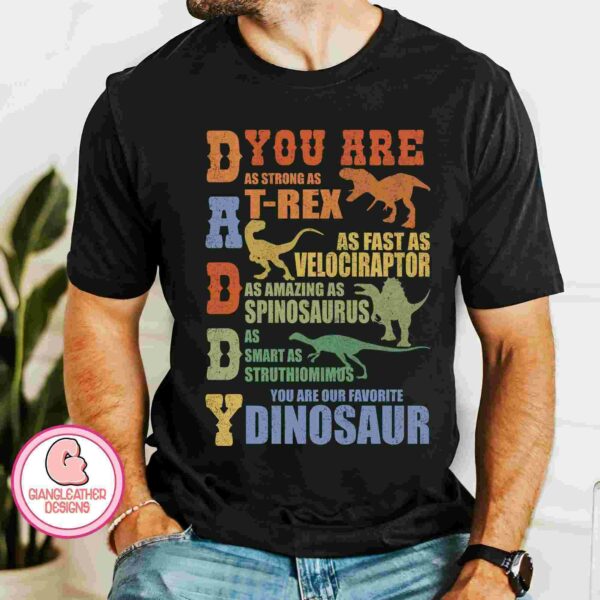 A person wears a black T-shirt with colorful text and dinosaur illustrations, stating "DADDY, You are as strong as T-Rex, as fast as Velociraptor, as amazing as Spinosaurus, as smart as Struthiomimus, you are our favorite dinosaur.