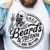 A person wearing a denim jacket and a white T-shirt displaying a skeleton with tattoos and the text "Dads with Beards & Tattoos Are Better.