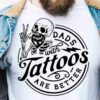 A person wearing a denim jacket and a white t-shirt with a graphic of a skeleton making a hand gesture, accompanied by the text "Dads with Tattoos Are Better.