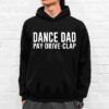 A person is wearing a black hoodie with white text that says, "DANCE DAD PAY·DRIVE·CLAP" standing against a white brick wall.