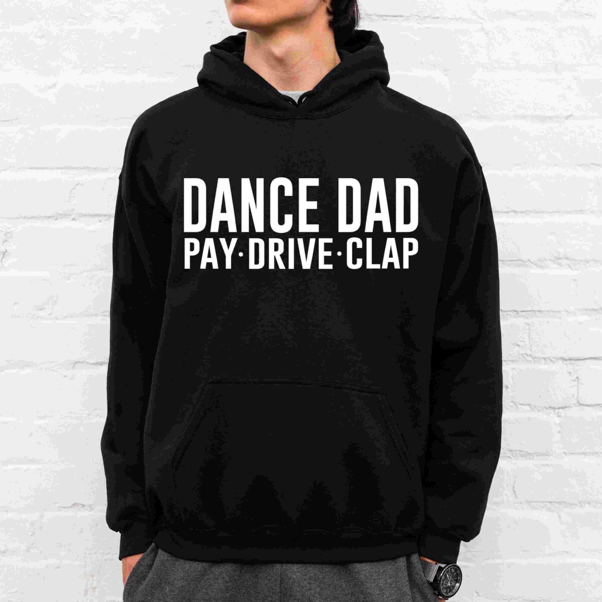 A person is wearing a black hoodie with white text that says, "DANCE DAD PAY·DRIVE·CLAP" standing against a white brick wall.