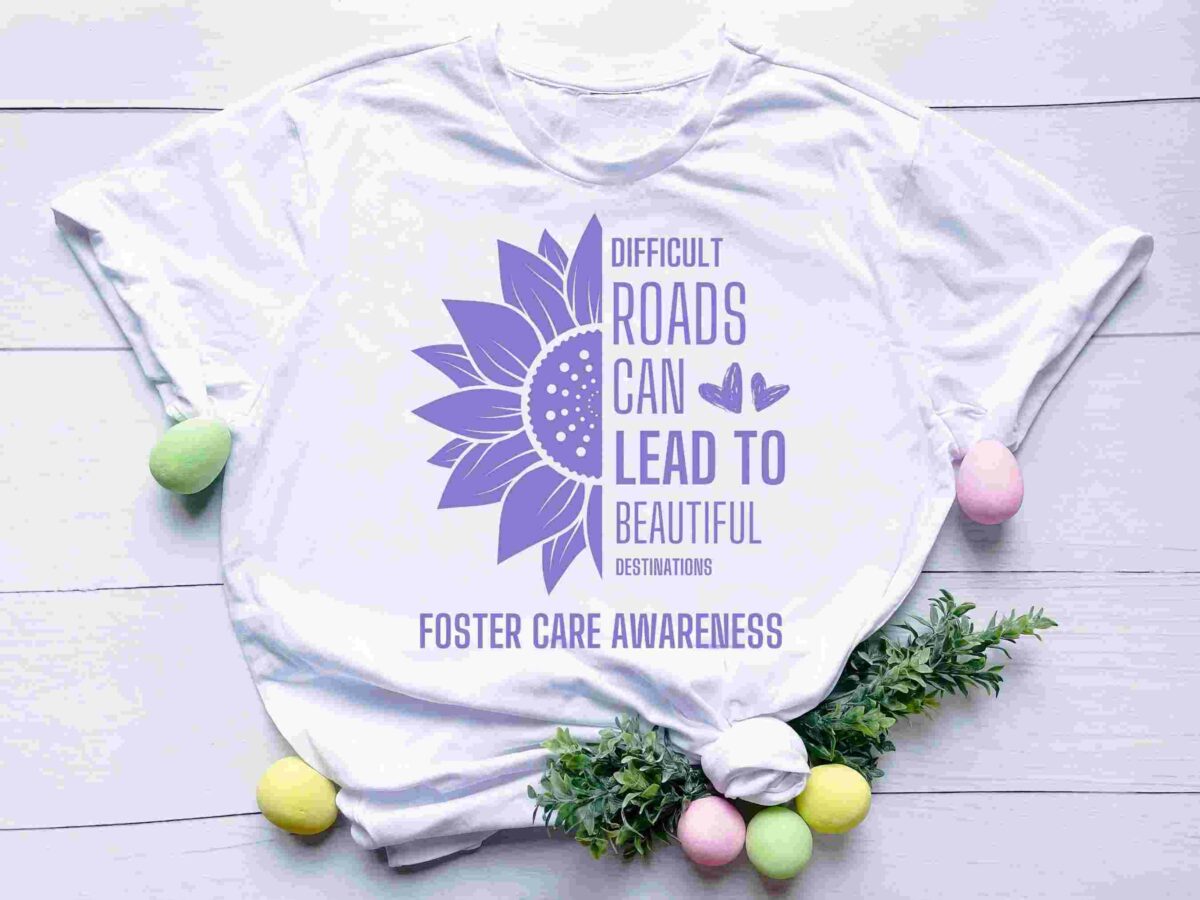 A white T-shirt with a purple sunflower graphic and the text "Difficult roads can lead to beautiful destinations" and "Foster Care Awareness." The shirt is on a white wooden background, decorated with pastel-colored eggs and green foliage.