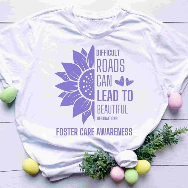 A white T-shirt with a purple sunflower graphic and the text "Difficult roads can lead to beautiful destinations" and "Foster Care Awareness." The shirt is on a white wooden background, decorated with pastel-colored eggs and green foliage.