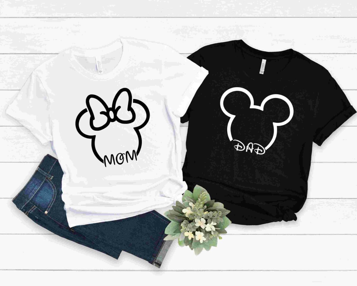 Two t-shirts on a wooden surface. The white shirt features a Minnie Mouse head with "MOM" written below, while the black shirt has a Mickey Mouse head with "DAD" written below. A pair of jeans and flowers are nearby.