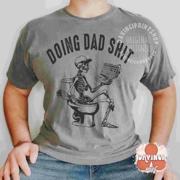 A person wearing a grey t-shirt with a graphic of a skeleton sitting on a toilet, holding a newspaper. The text on the shirt reads "DOING DAD SH*T." The image has a watermark for "DAVINCI PRINTSHOP." The person is standing against a plain background.