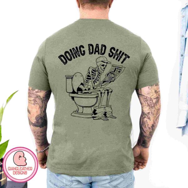 A man with tattoos is wearing a green t-shirt that features a skeleton sitting on a toilet and reading a newspaper with the text "DOING DAD SHIT" on the back.