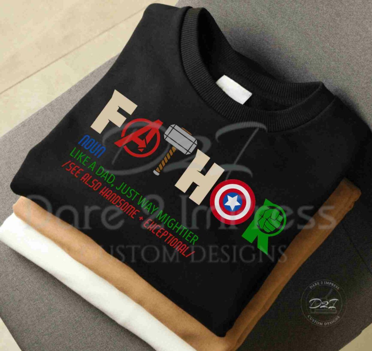 A neatly folded black sweatshirt with "FATHER" written in colorful superhero-themed letters and a playful definition below it, featuring references to superheroes and qualities such as "awesome" and "exceptional.