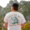 A person wearing a white shirt with a "Follow Me & I Will Make You Fishers of Men" graphic stands facing a mountainous landscape.